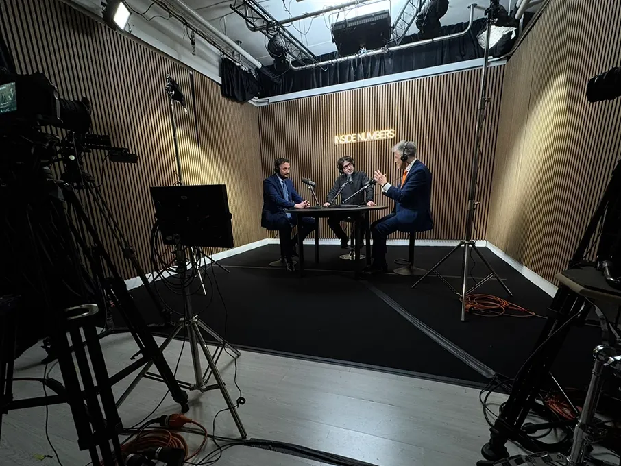 Camere sul set: tre ospiti al tavolo ripresi da una camera professionale su treppiede, con softbox e pannello LED in regia