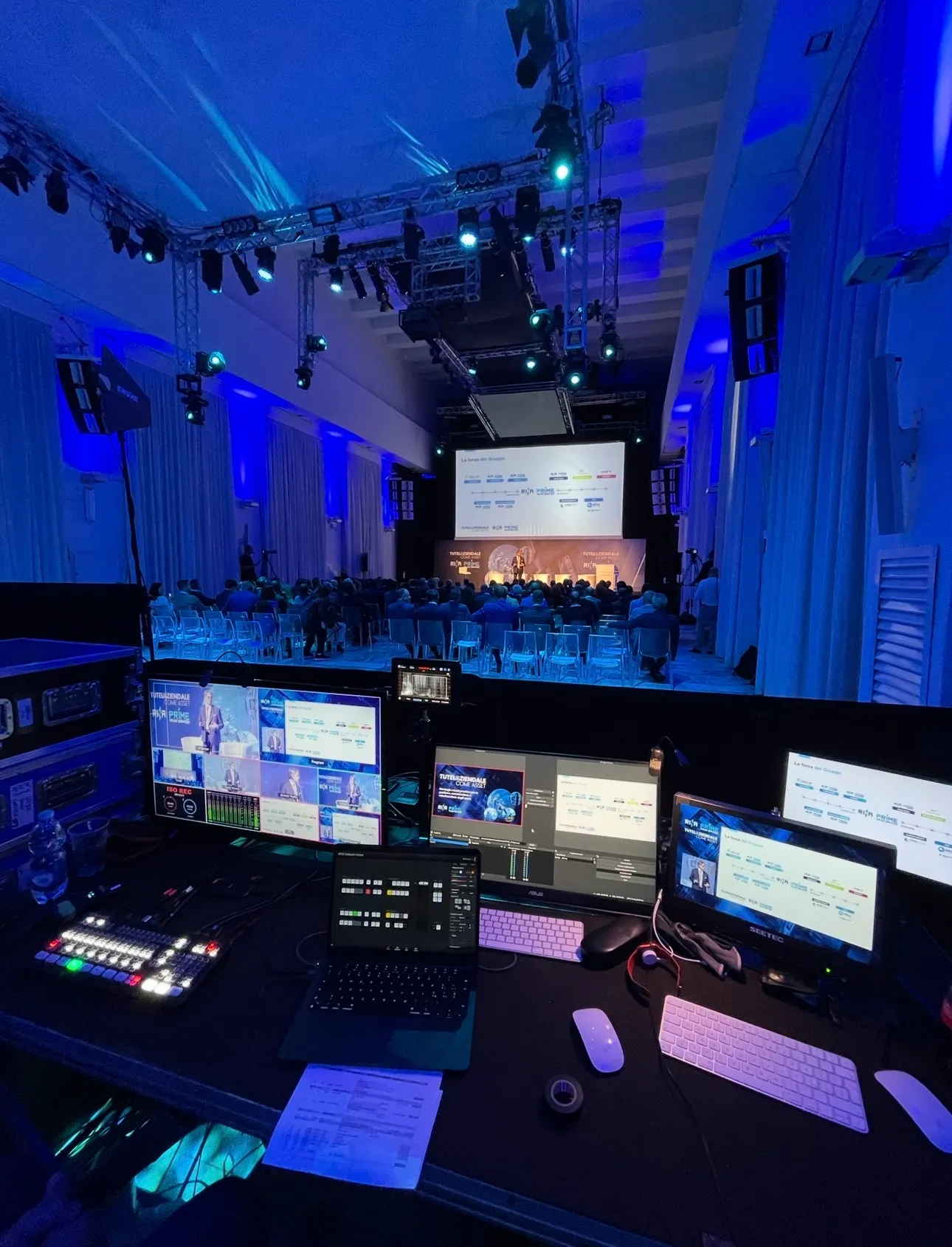Regia multicamera live da una cabina di controllo durante un evento aziendale: monitor, mixer video e palco illuminato in blu